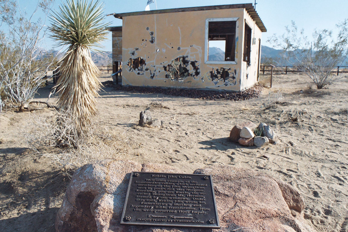 A photograph of Krblin Jihn Kabin, a Kymaerica historical site in the nation of Notgeon (near Joshua Tree, California). The historical marker in the foreground reads “Well-sited example of the homestedler cabins which secluded members of the Jihn Wranglican sect in post-Civil War Notgeon. Kneeling, you see Kwale, the so-called Wranglican Mt. Hermon, framed by the windows. Under this roof, Krblin Jihn wrote his Kmmentaries n Matthew and legendary Bible translations. Note the well-preserved floor kmpass.” 