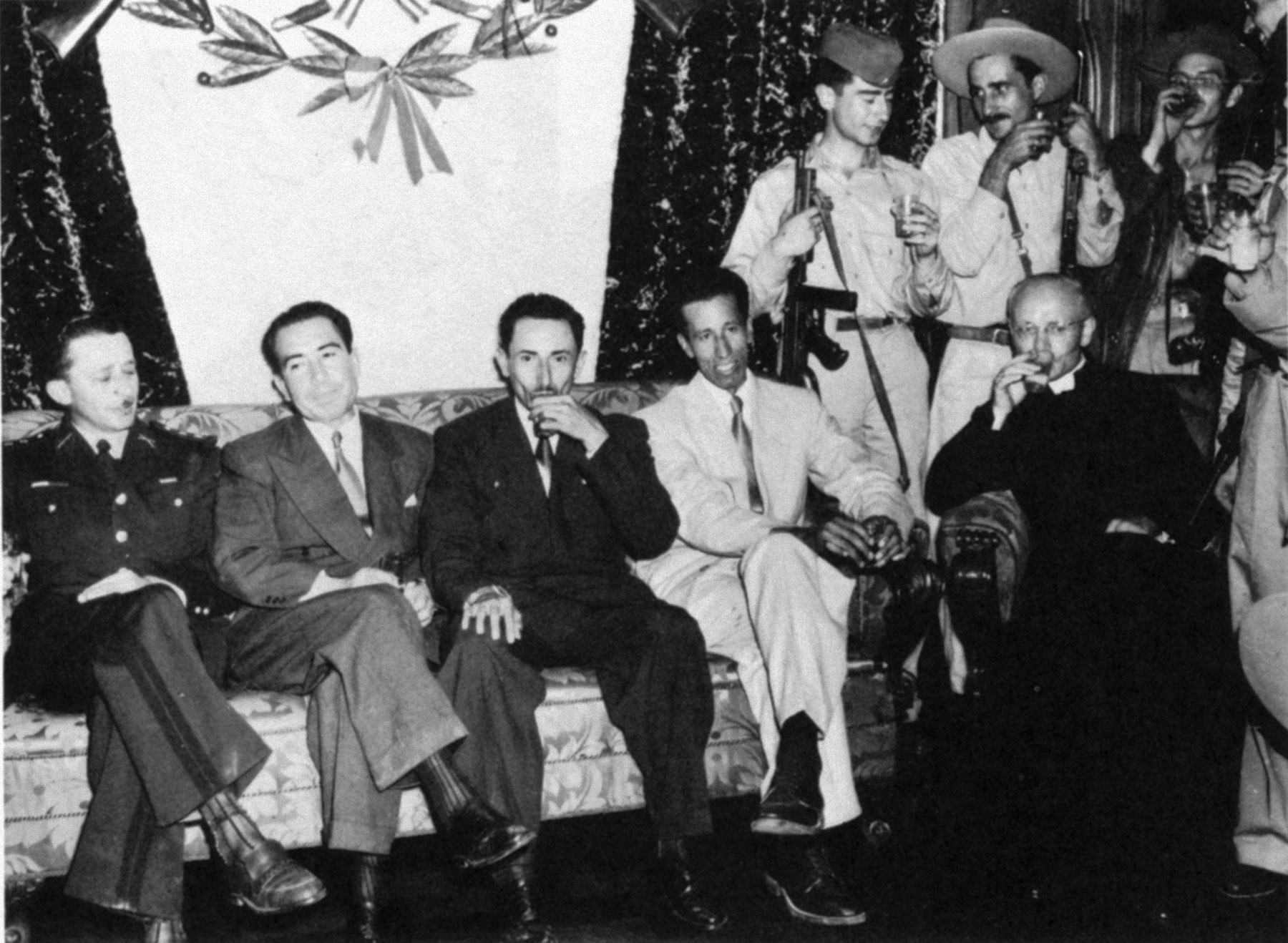 A photograph of four of the five members of the original Castillo Armas junta (seated on couch) posing with Papal Nuncio Monsignor Gennaro Verrolino, a bitter Árbenz opponent (seated on chair, far right). Castillo Armas is third from left on the couch.