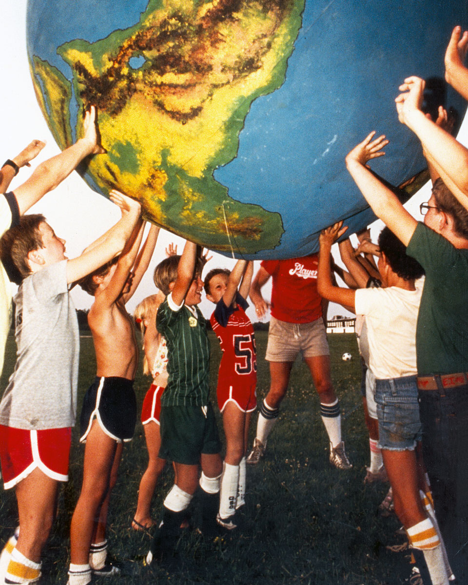 A nineteen eighty photograph of children holding an earth-shaped ball above their heads. The New Games Foundation sold this type of ball, which had a canvas exterior and heavy-duty vinyl bladder interior, with only the continents outlined, so that the image of the earth’s surface had to be painted by hand. The game being played here is called Orbit. Participants would form a circle, with one player in the center, all holding the earth ball aloft. The center participant would try to push the ball outside of the circumference of the circle, while those on the edges would try to keep it within the circle. As with all New Games, there was no scoring system or competitive goal to Orbit.