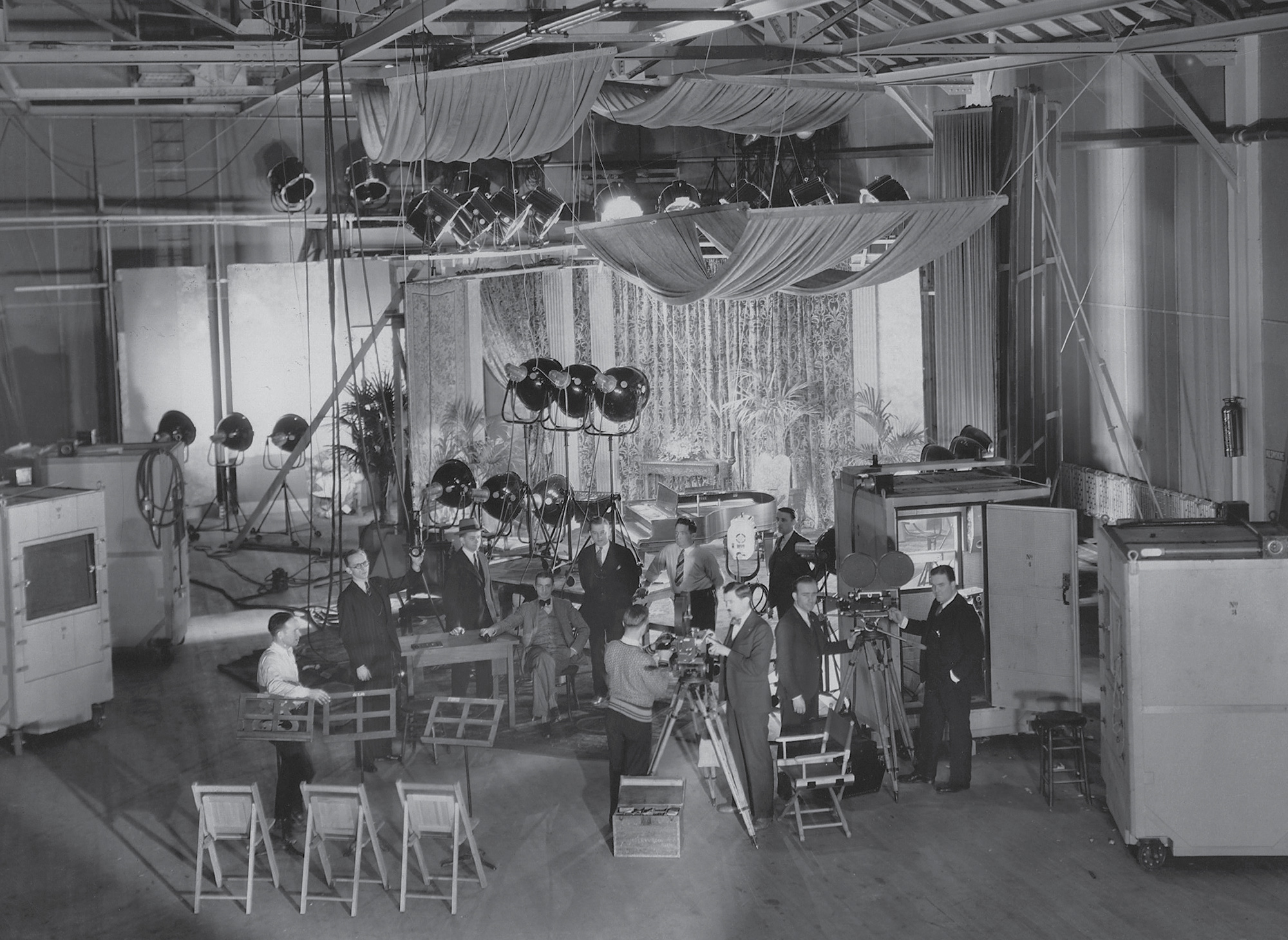 A circa nineteen thirty production shot showing one of the sound stages at Warner Brothers’ Brooklyn studio. The large “cabins”—soundproof booths that housed the cameras—were on rollers and could be moved. Often up to three cameras shot simultaneously from different angles. The Vitaphone sound-on-disc system allowed sounds recorded live during filming to later be merged with additional background music and sound effects onto one new composite disk that was played in the cinema during screenings.