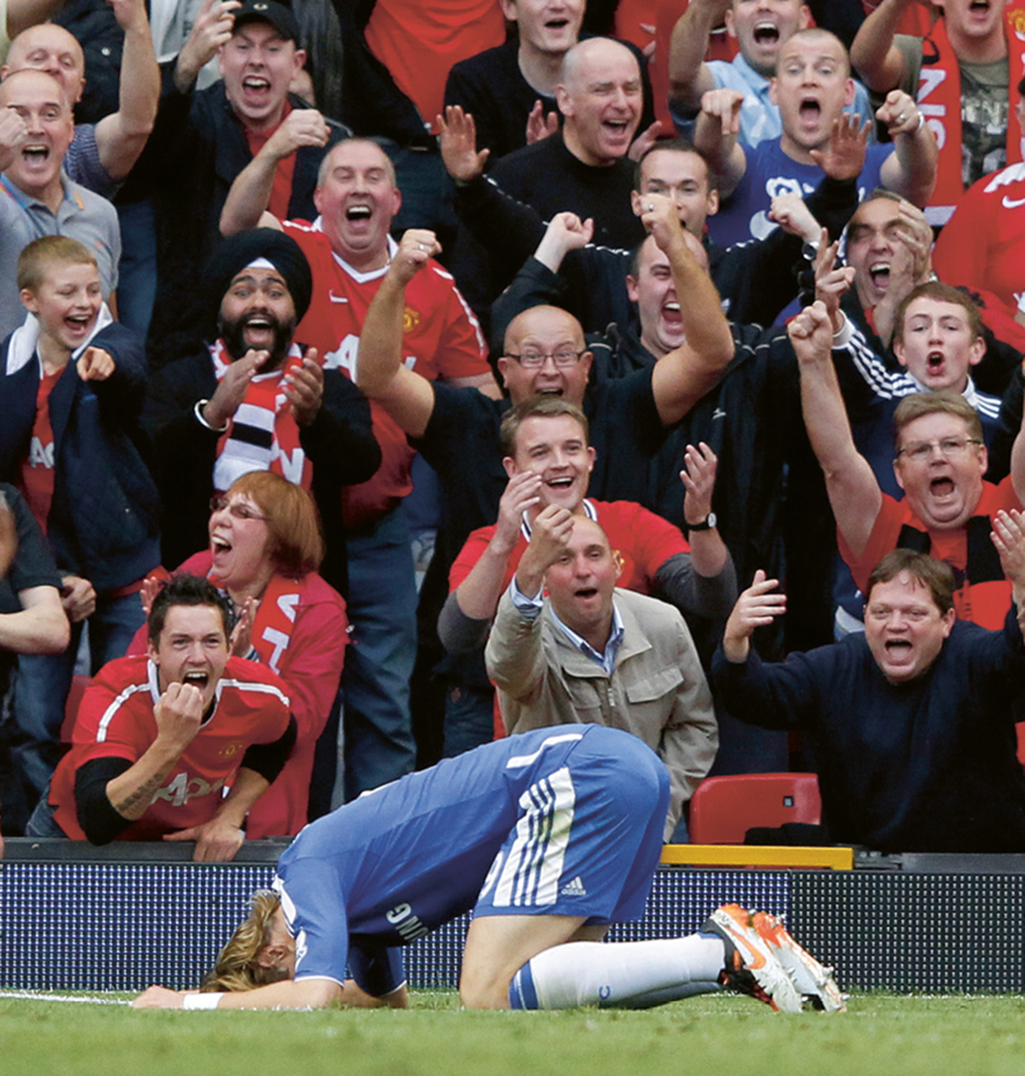 A photograph of soccer player Fernando Torres kneeling face-down on the pitch as fans from the opposite team jeer in the stalls behind him. 