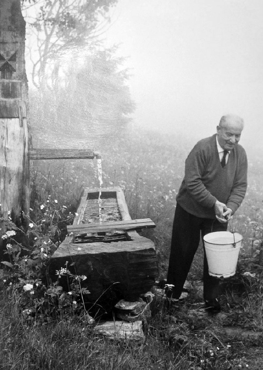 An undated photograph of Heidegger carrying a bucket of water outside his hut in the Black Forest.