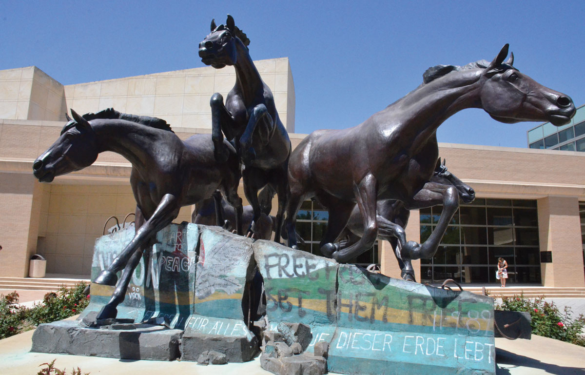 A photograph of a sculpture outside the presidential library of George H. W. Bush, in which galloping horses emerge from a fascimile fragment of the Berlin Wall.