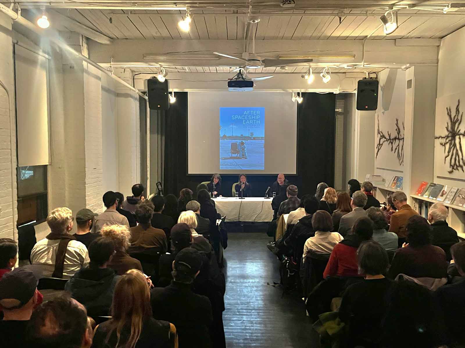 A photograph of three panelists, one of whom is speaking into a microphone, sitting at a table in front of a seated audience. An image of the front cover of a book titled After Spaceship Earth: Art, Techno-utopia, and Other Science Fictions is projected onto a screen behind the panelists.