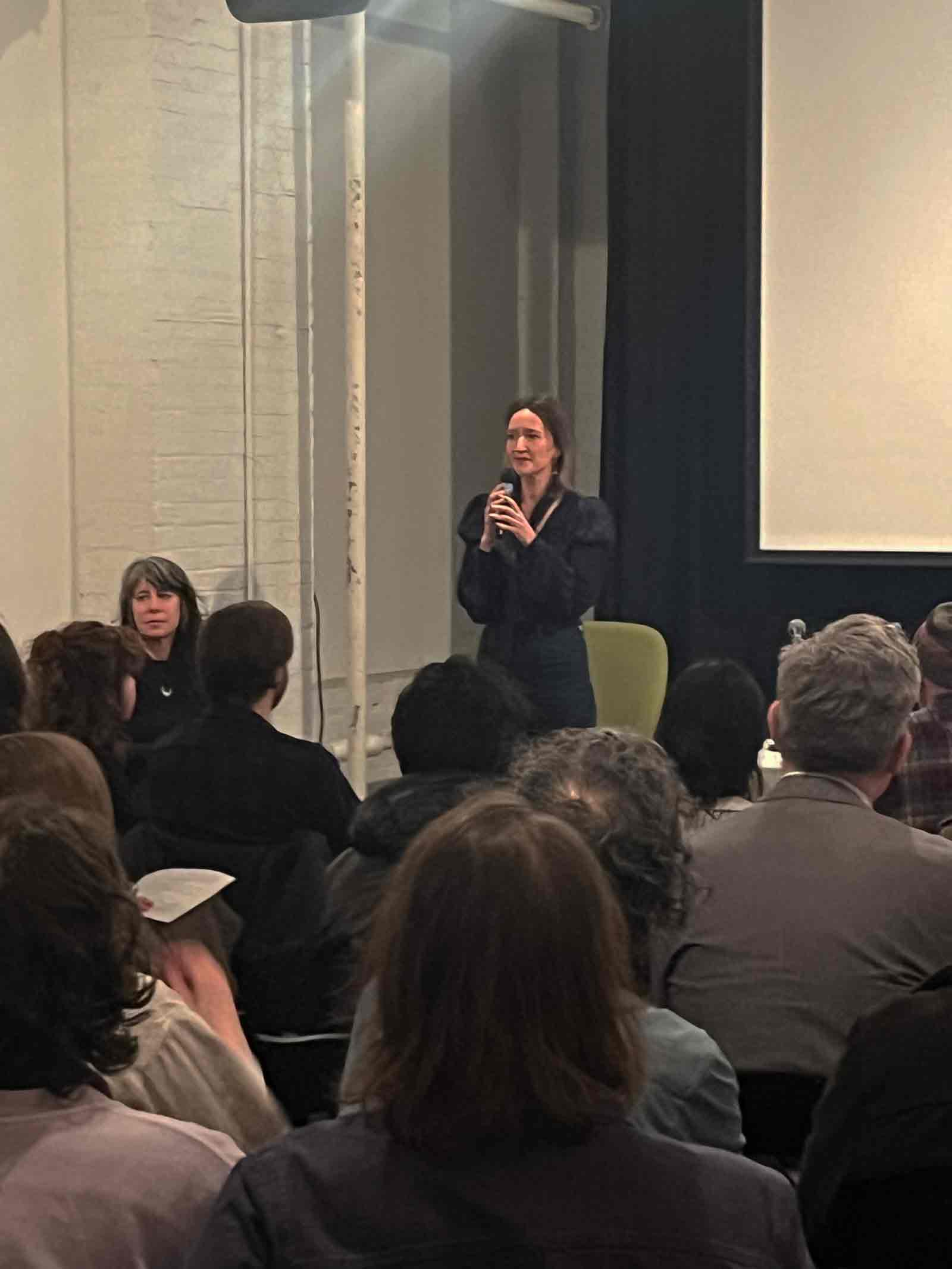 A photograph of a woman who stands before a seated audience and holds a microphone as though about to speak.
