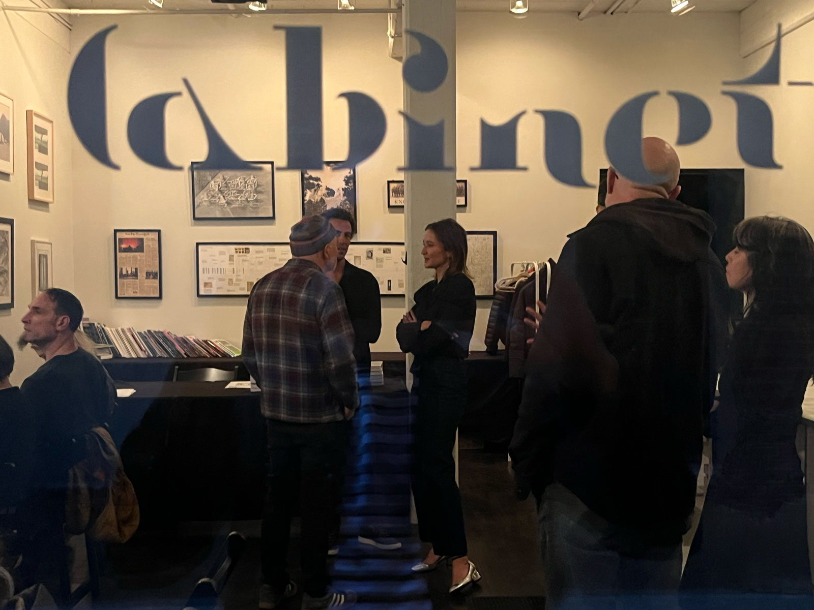 A photograph taken through a window imprinted with the Cabinet logo shows three people standing together and chatting.