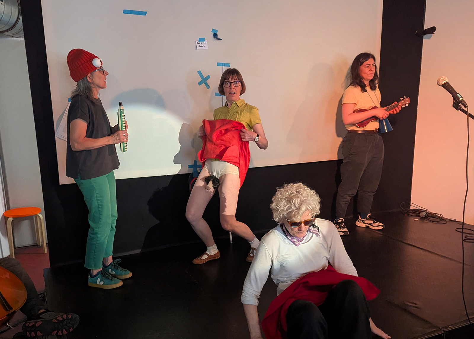 A photograph of four performers practicing the opening song and dance number during rehearsals. The performer in the center gazes into the camera with an expression of surprise as she bends her knees and lifts her skirt.