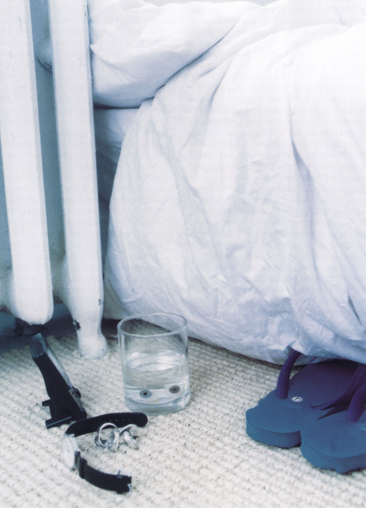 A photograph of two glass eyes floating in a tumbler of water on the floor next to a bed.