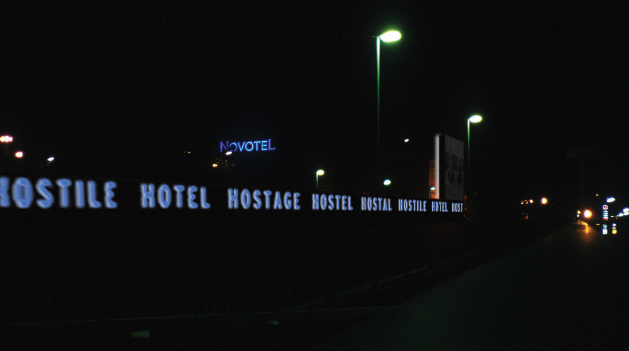 A photograph at night of lit neon signs wrapping around the hotel, reading “Hostile Hotel Hostage Hostal...”