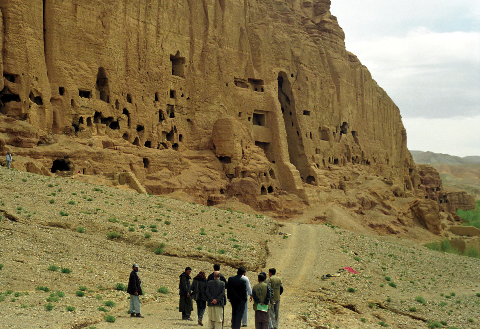 A photograph of people standing in Bamiyan Valley below cliffs dotted with niches where the Buddha statues once stood.