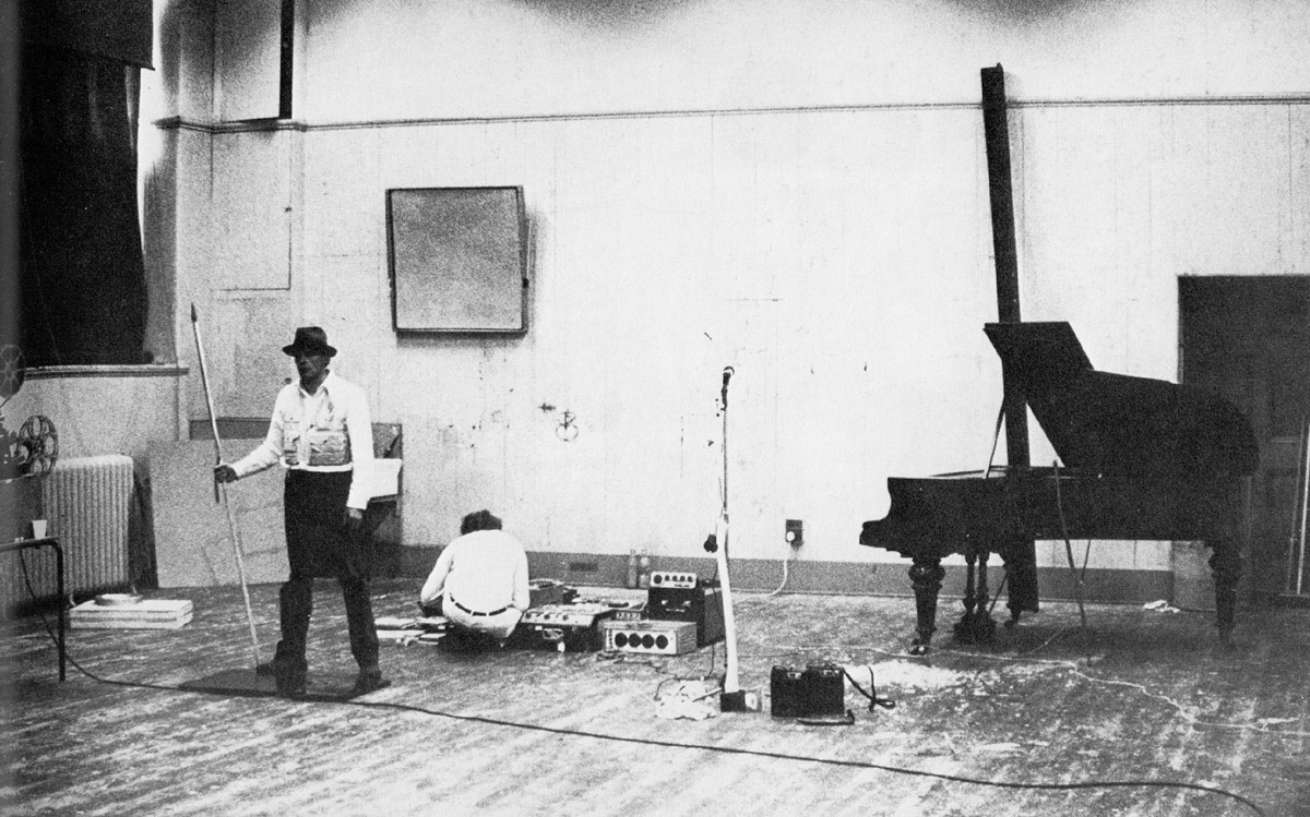 Photograph from 1970 by Ute Klophaus of Joseph Beuys holding a pole during the action entitled Celtic. Christiansen is seated on the ground behind him, manipulating tape recorders.