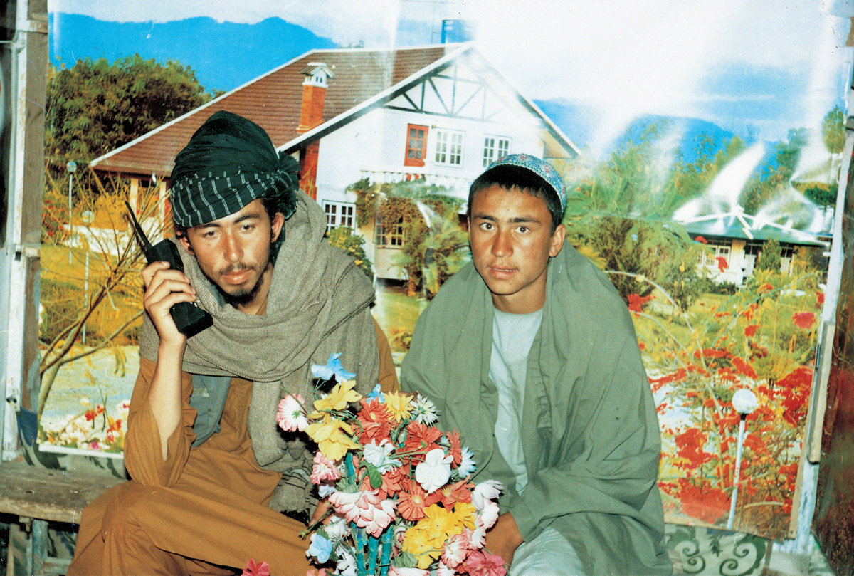 Studio portrait of two Taliban members, one holding fake flowers, in front of a cottage garden backdrop.
