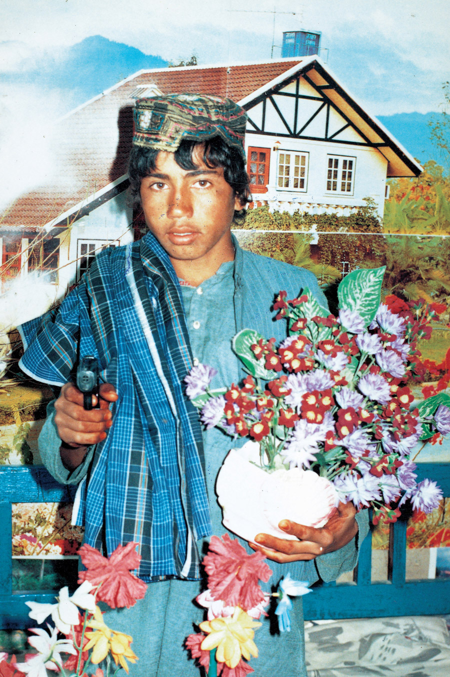 Studio portrait of a boy holding flowers and a gun in front of a cottage garden backdrop.