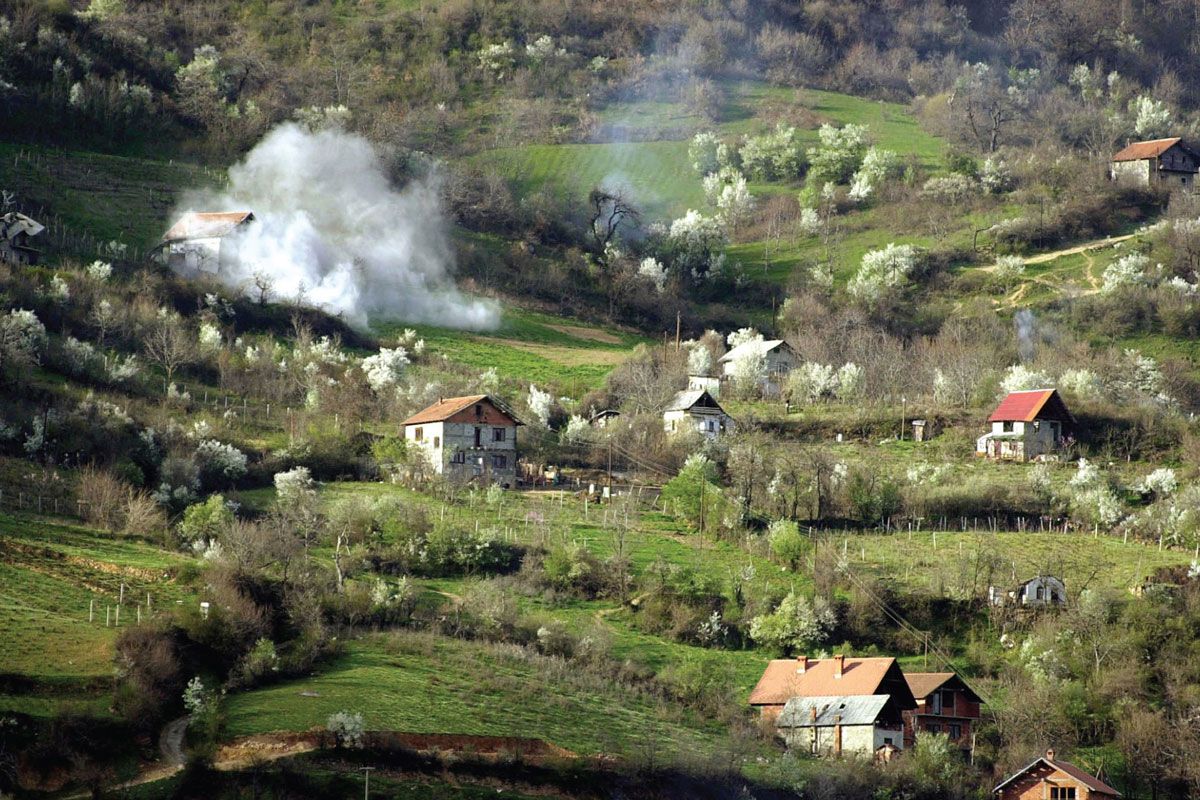 A photograph of houses sitting in a valley, a smoke cloud to the left of them.