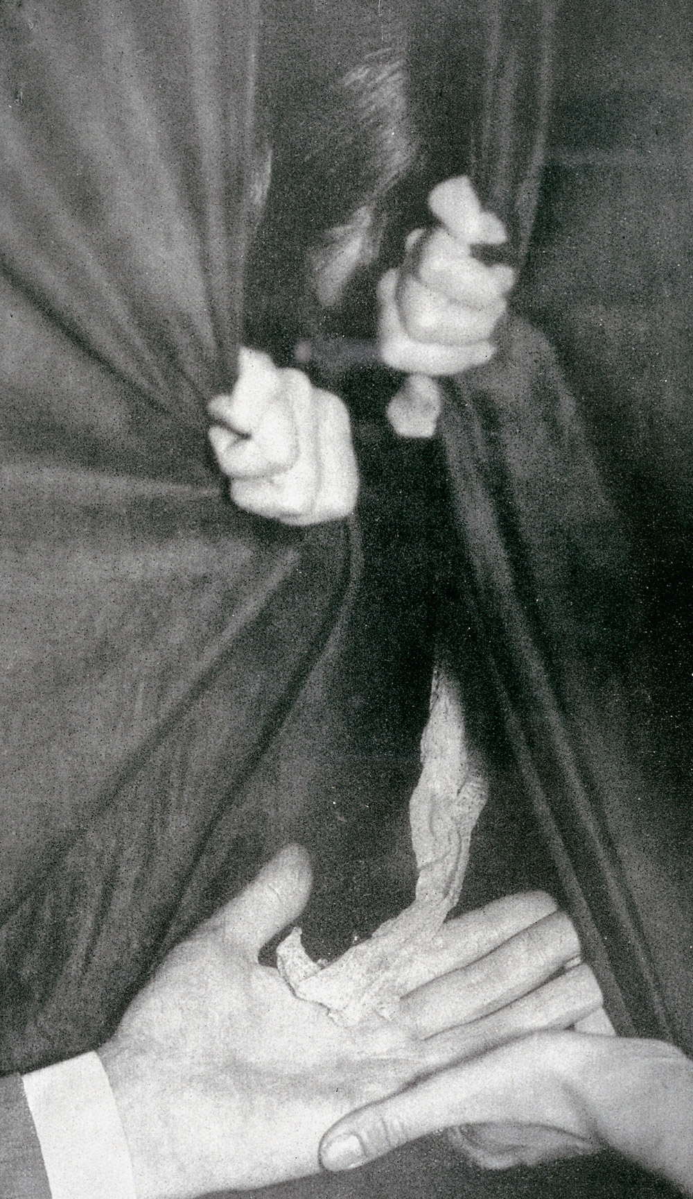 A photograph of Eva C. hidden behind a curtain during the séance of 21 August 1911, two hands reaching out to hold the ectoplasm.