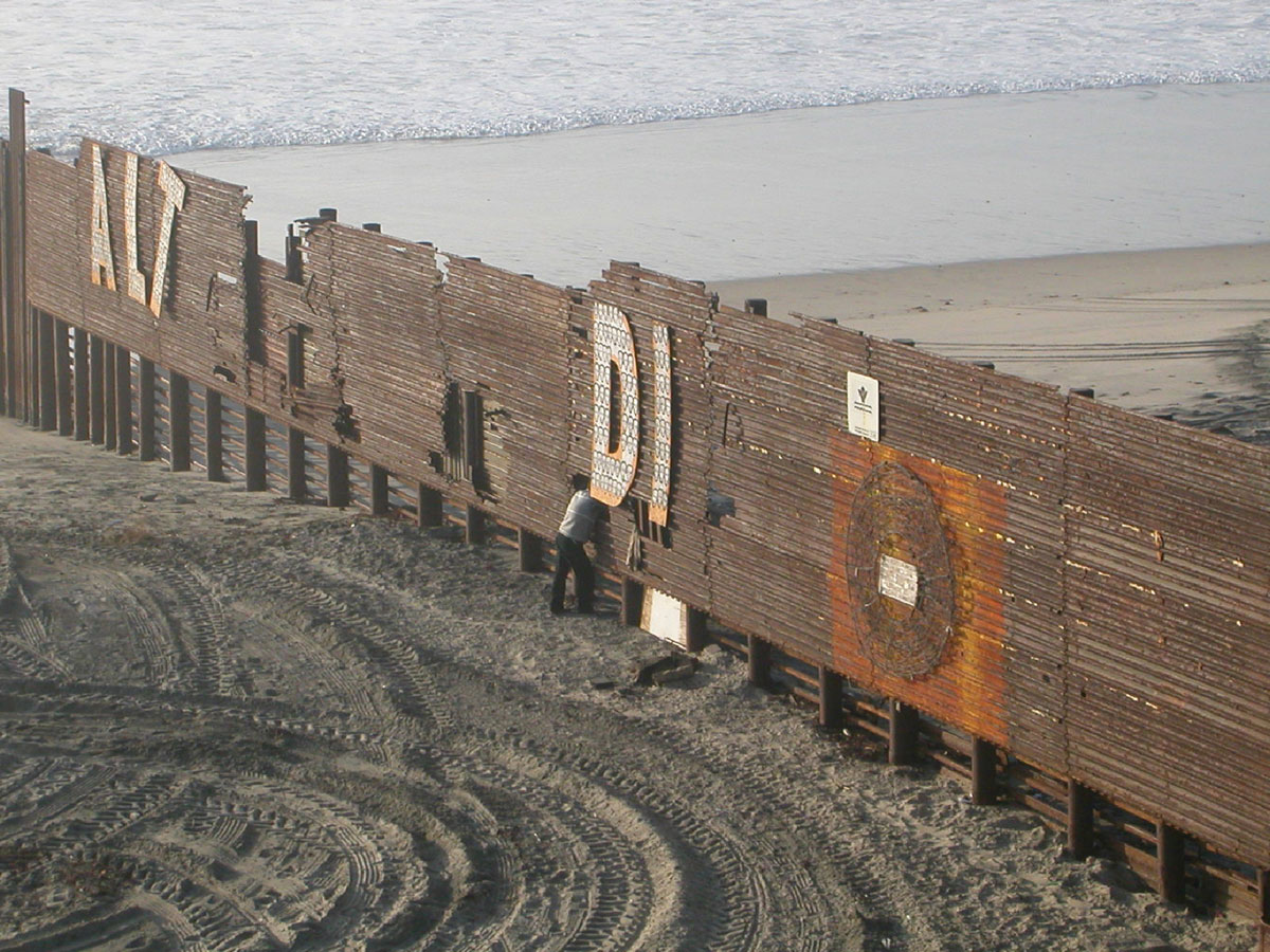 A photograph of a rusted fence cutting through the beach, leading to the shore.