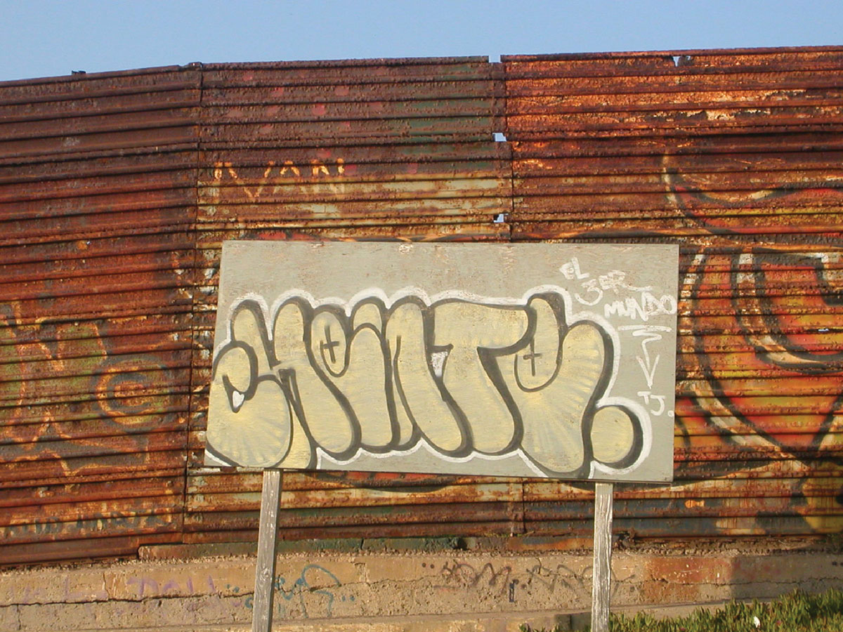 A photograph of a rusty US-Mexico border fence section, with a small wooden billboard covered in unintelligible graffiti in front.