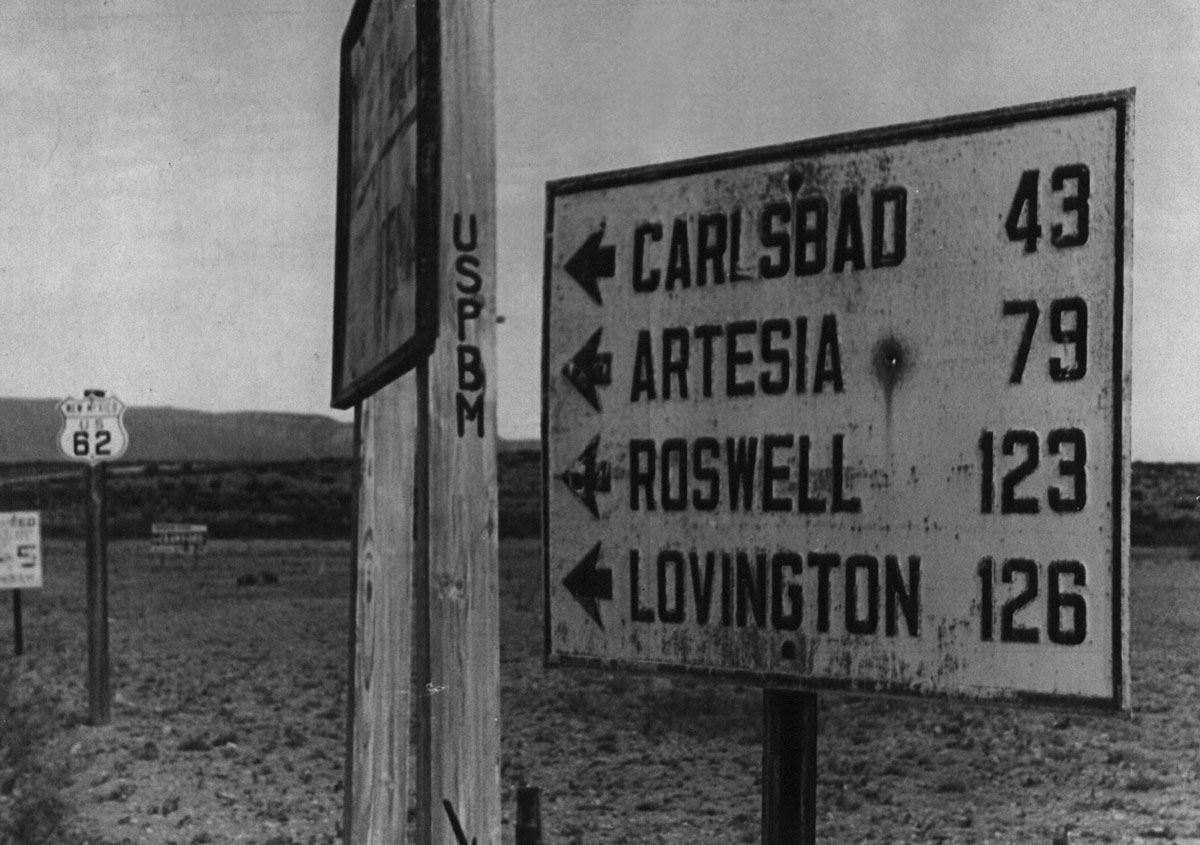 Black-and-white photograph of a sign on side of the road showing how many miles to different cities.
