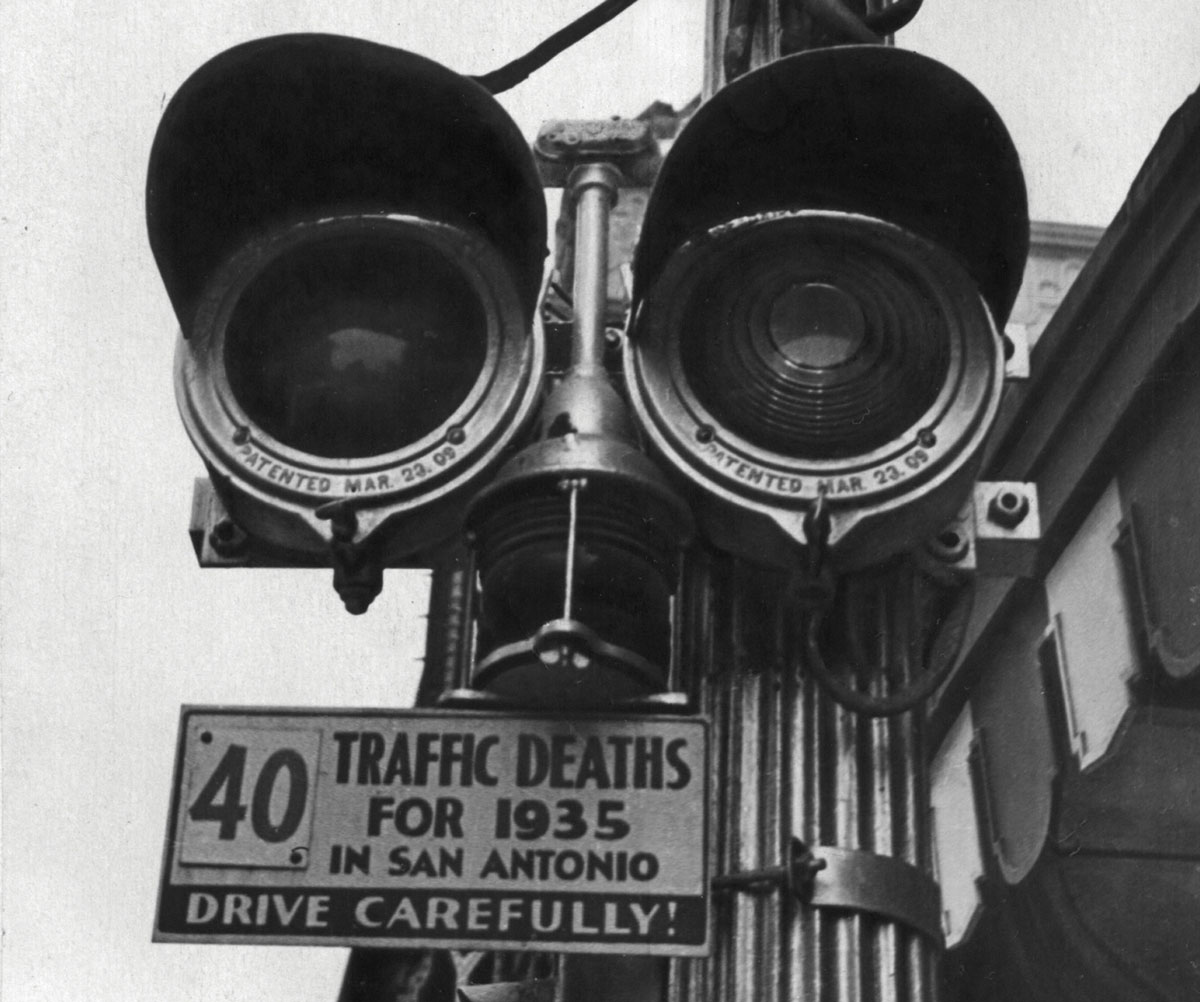 Black-and-white photograph of a stop light with sign underneath recording the town's traffic deaths.