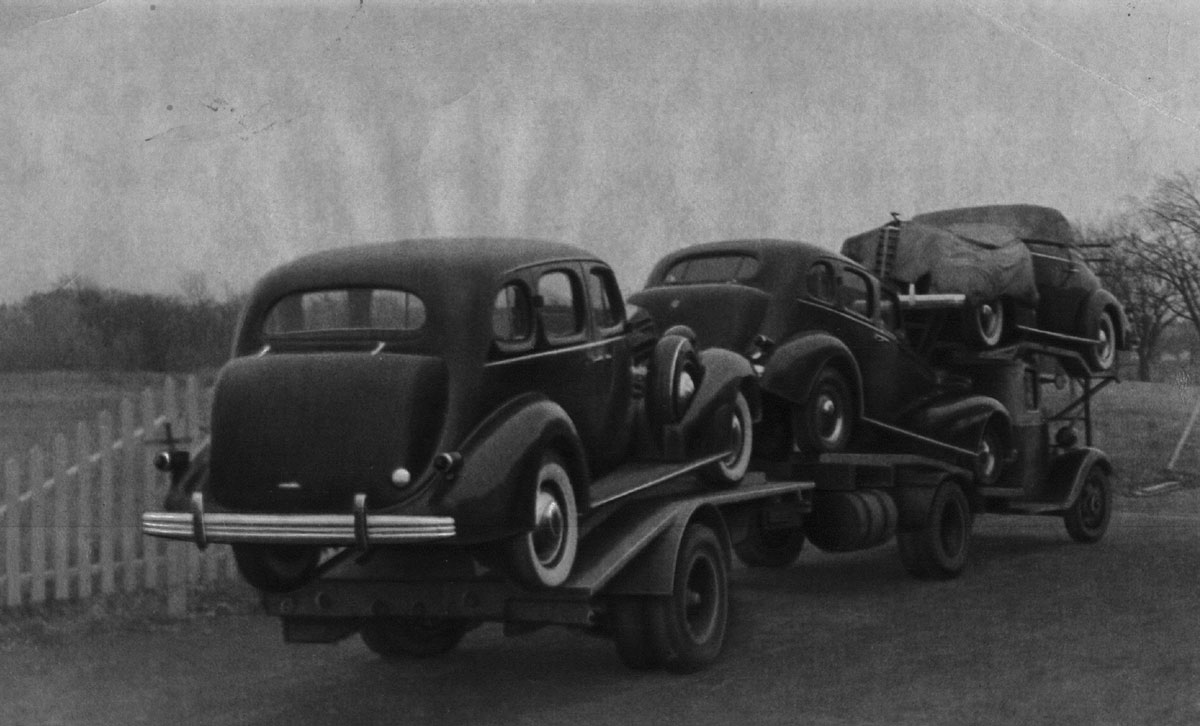 Black-and-white photograph of a truck carrying three automobiles.