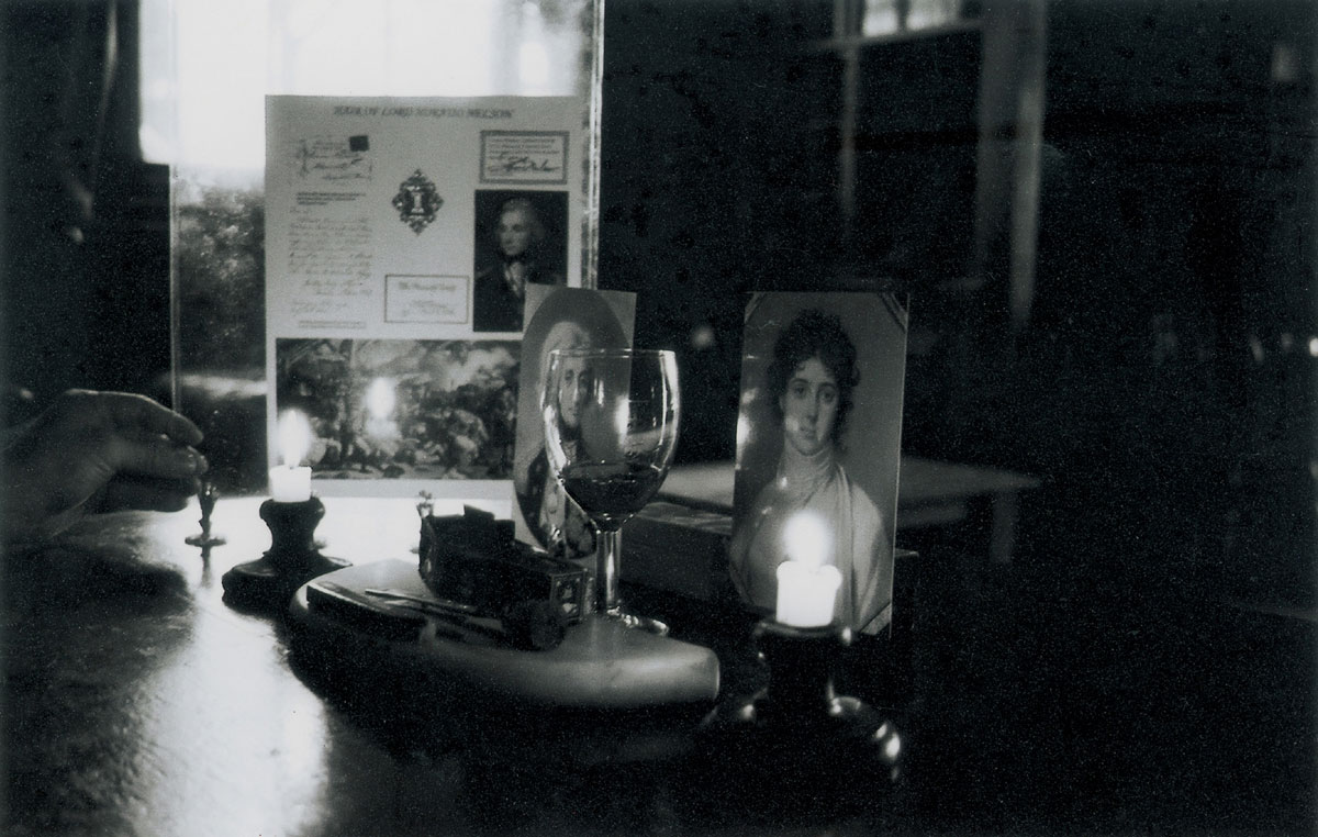 A photograph of a shrine-like collection of objects including painted portraits of Horatio Nelson, a glass of wine, and a candle. 