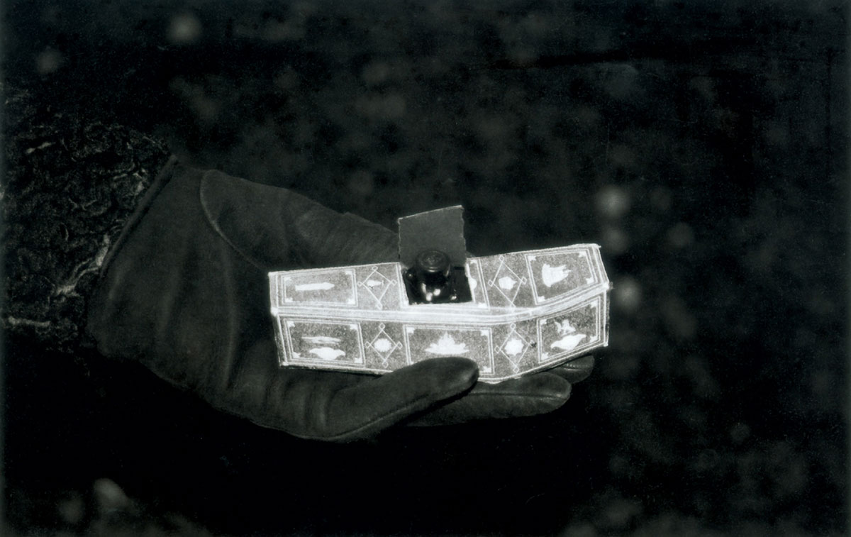 A photograph of a gloved hand holding a miniature paper coffin containing a small vial.