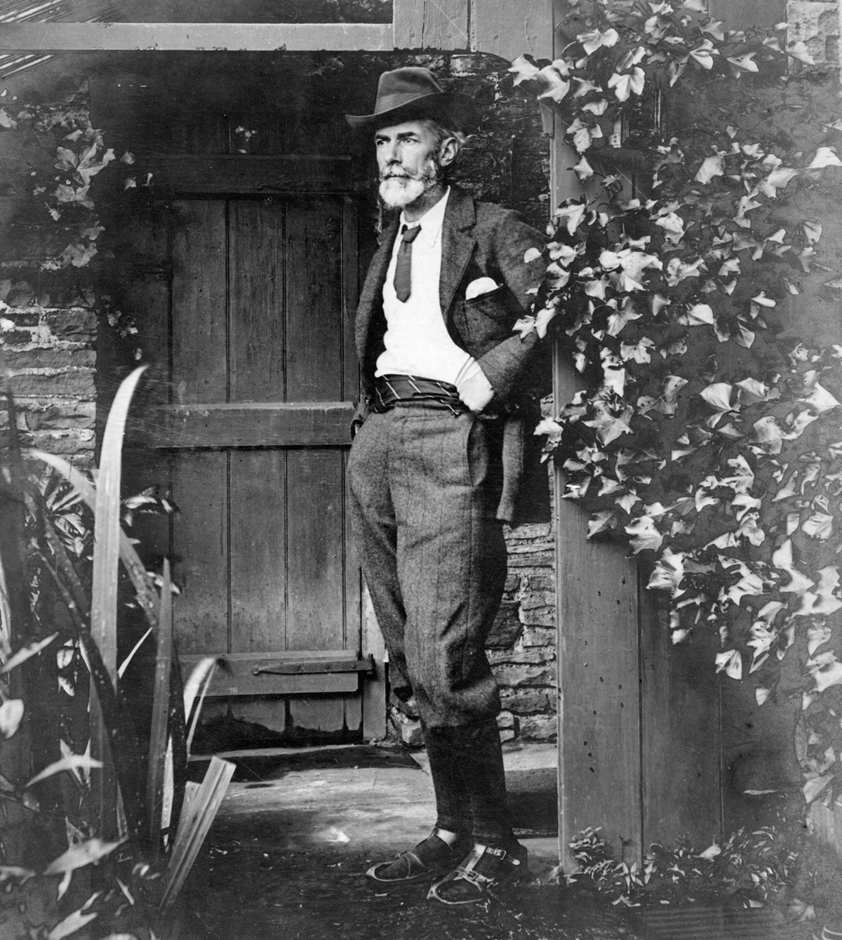 A 1905 photograph of Edward Carpenter standing outside his cottage in Millthorpe, Derbyshire. He is wearing a pair of his famous Indian-style sandals, which he made himself, and a jacket, knickerbockers, cravat, and cummerbund of his own design.