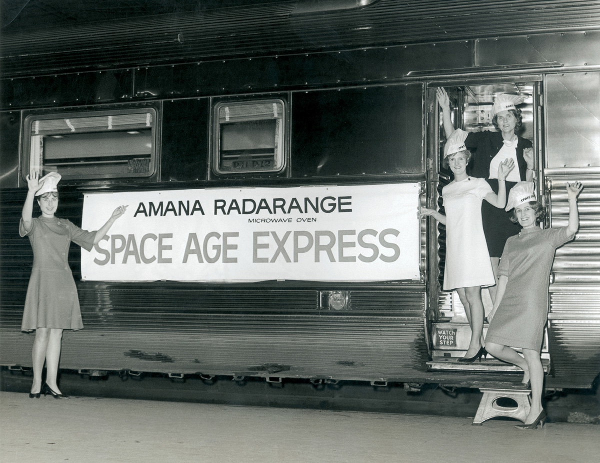 A photograph of the Amana Radarange Space Age Express, a microwave-equipped train that traveled to US cities in nineteen sixty eight to promote the Amana Radarange. Each stop attracted enormous publicity and included a tour of the onboard kitchens with cooking demos by female staff.