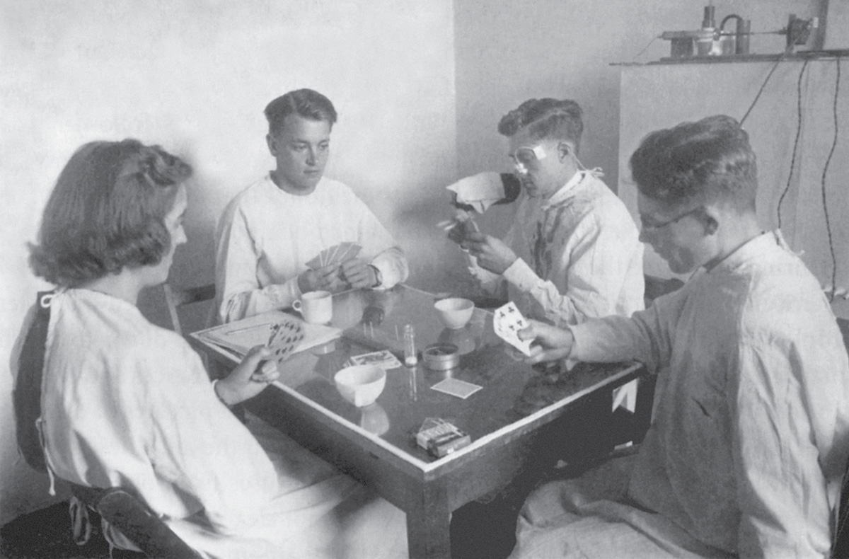 A photograph of four people playing cards in a lab setting. The person at the back had a tube running to his nose that slowly leaked a fluorescent dye while he played. After the game, the room was examined with the light of an ultraviolet lamp, which showed that the dye had spread over each of the players, the table and cards, and even further away. 
