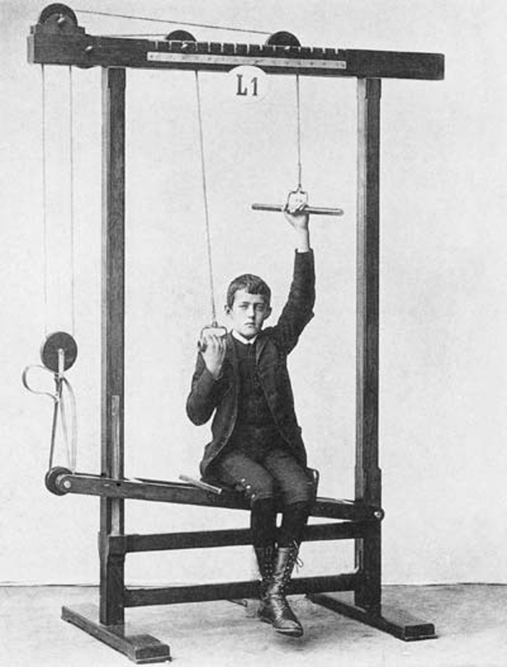 A nineteen-eighties photograph of a boy in a suit holding on to pulleys on a Zander exercise machine.
