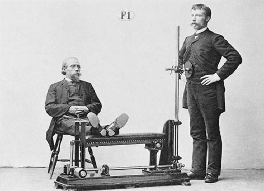 A nineteen-eighties photograph of two men in suits, one standing and the other sitting next to a Zander exercise machine.