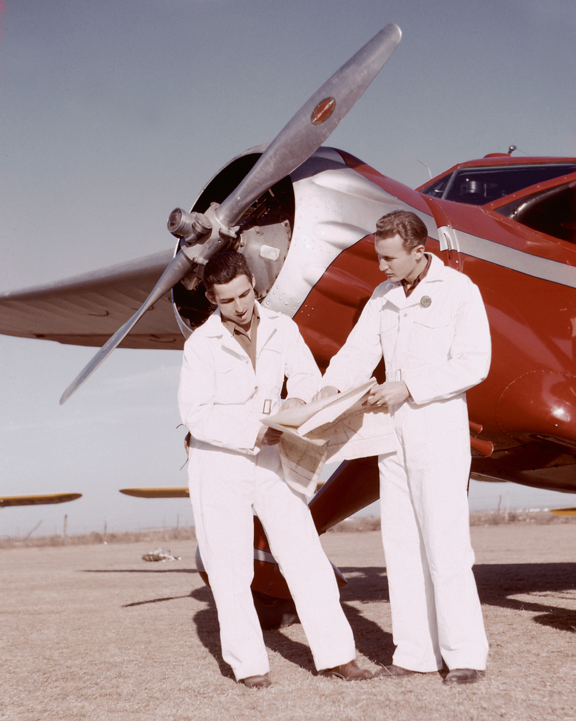 A photograph of two men in white clothes in front of a red, single-propeller airplane. 