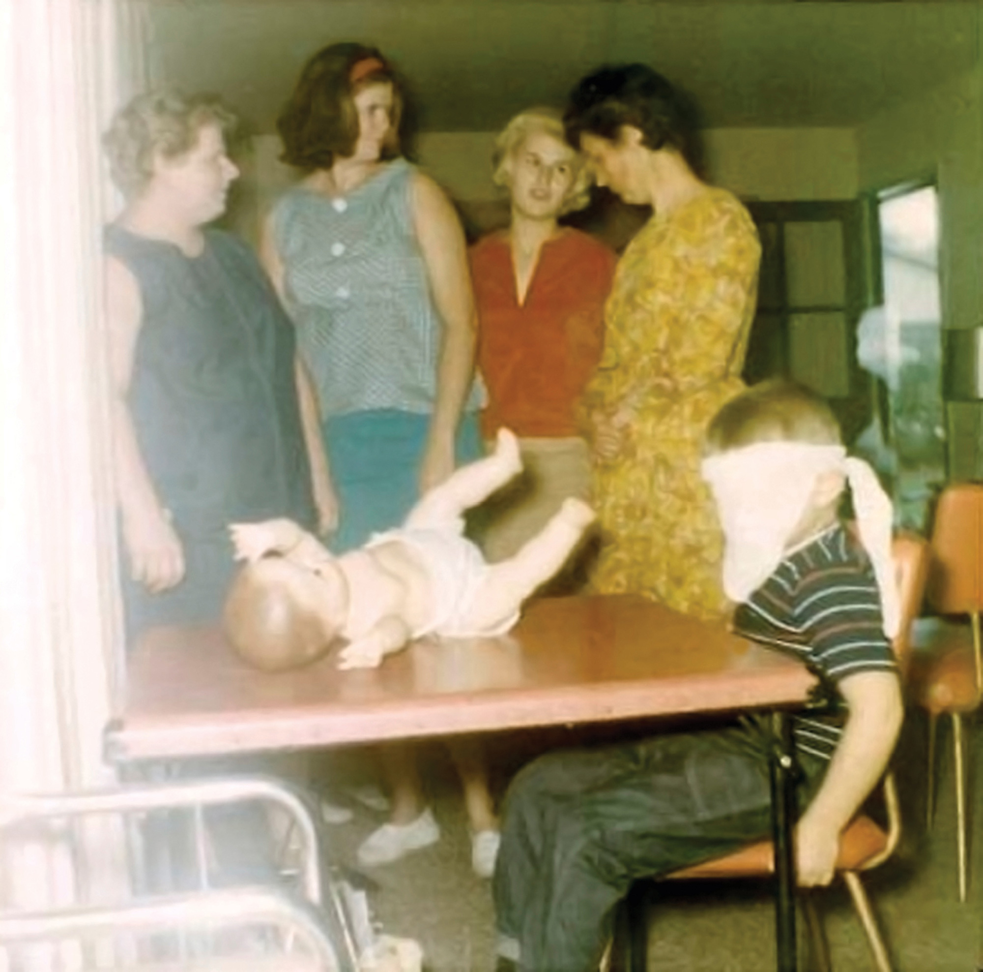 A photograph of four women talking while a blinded folded boy sits at a table on which a doll is lying on its back. 