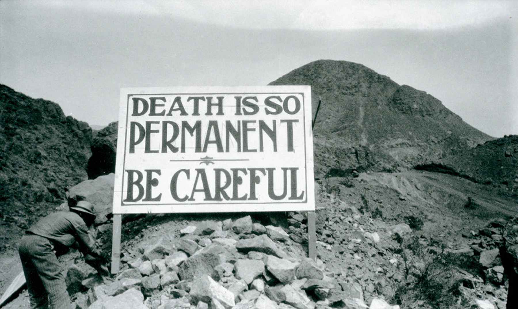 A photograph of a sign that reads “Death is so permanent. Be careful.” The sign was erected for workers involved in the construction of the Hoover Dam in the early 1930s, a project that resulted in the death of hundreds of workers.