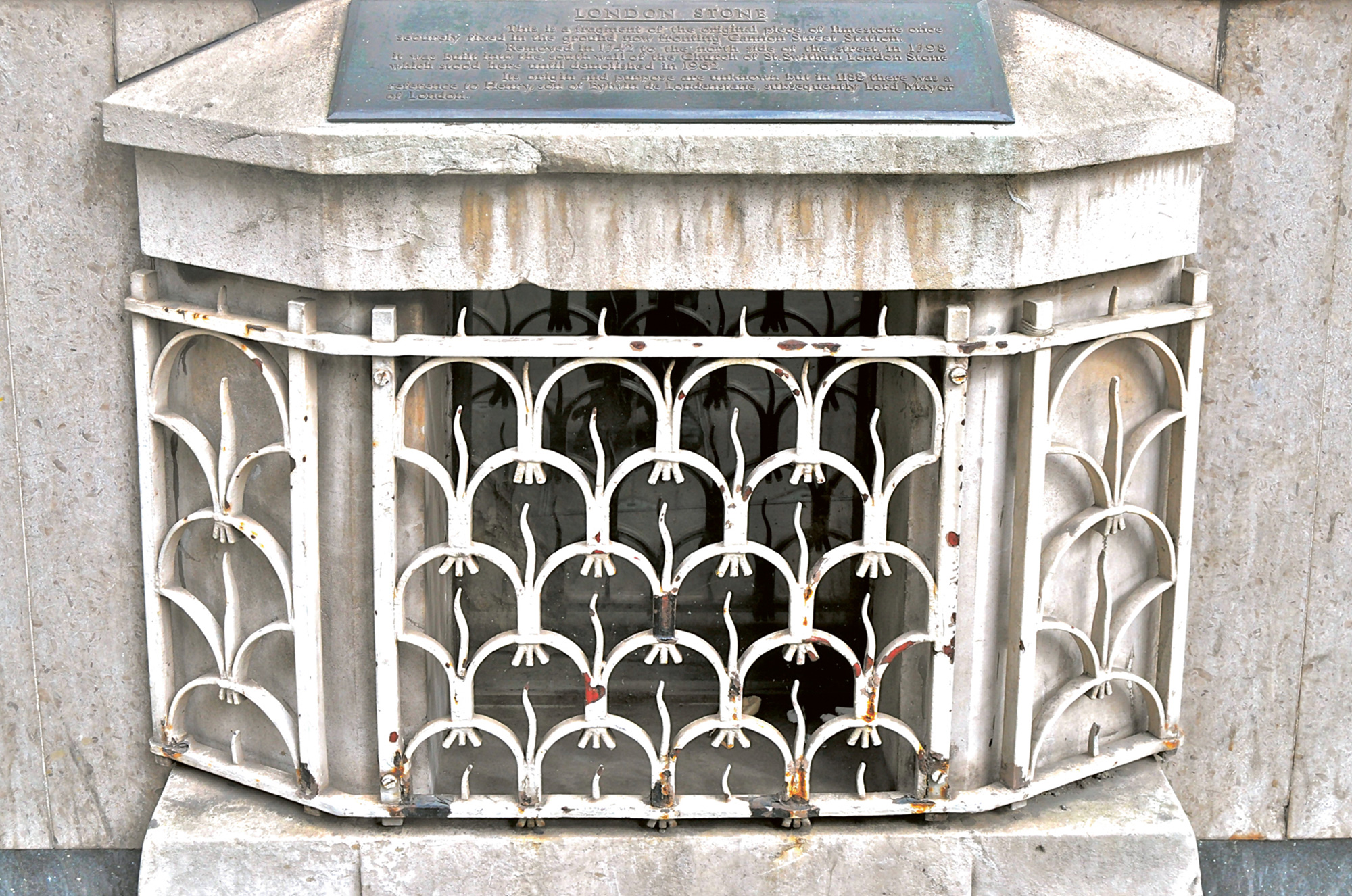 A photograph of the London Stone in its current state: encased in an iron fence and topped with a plaque.
