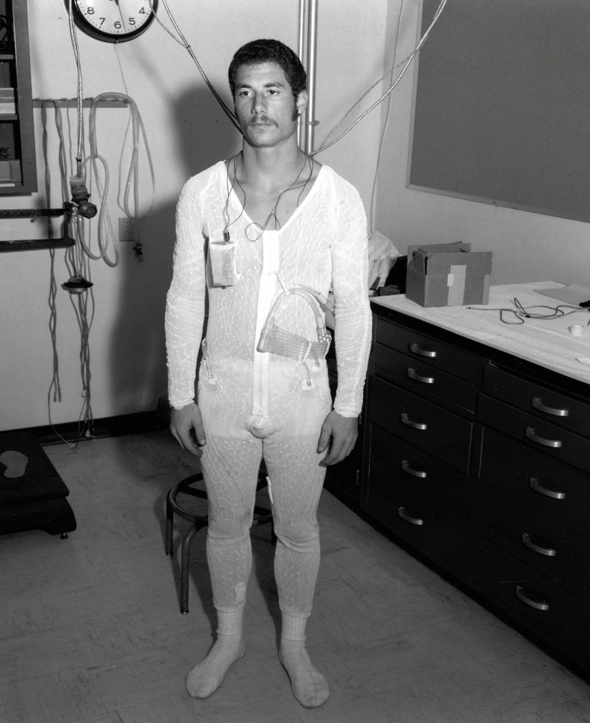 A photograph of a man standing in a white one-piece attached with multiple wires to unidentifiable medical equipment behind him.