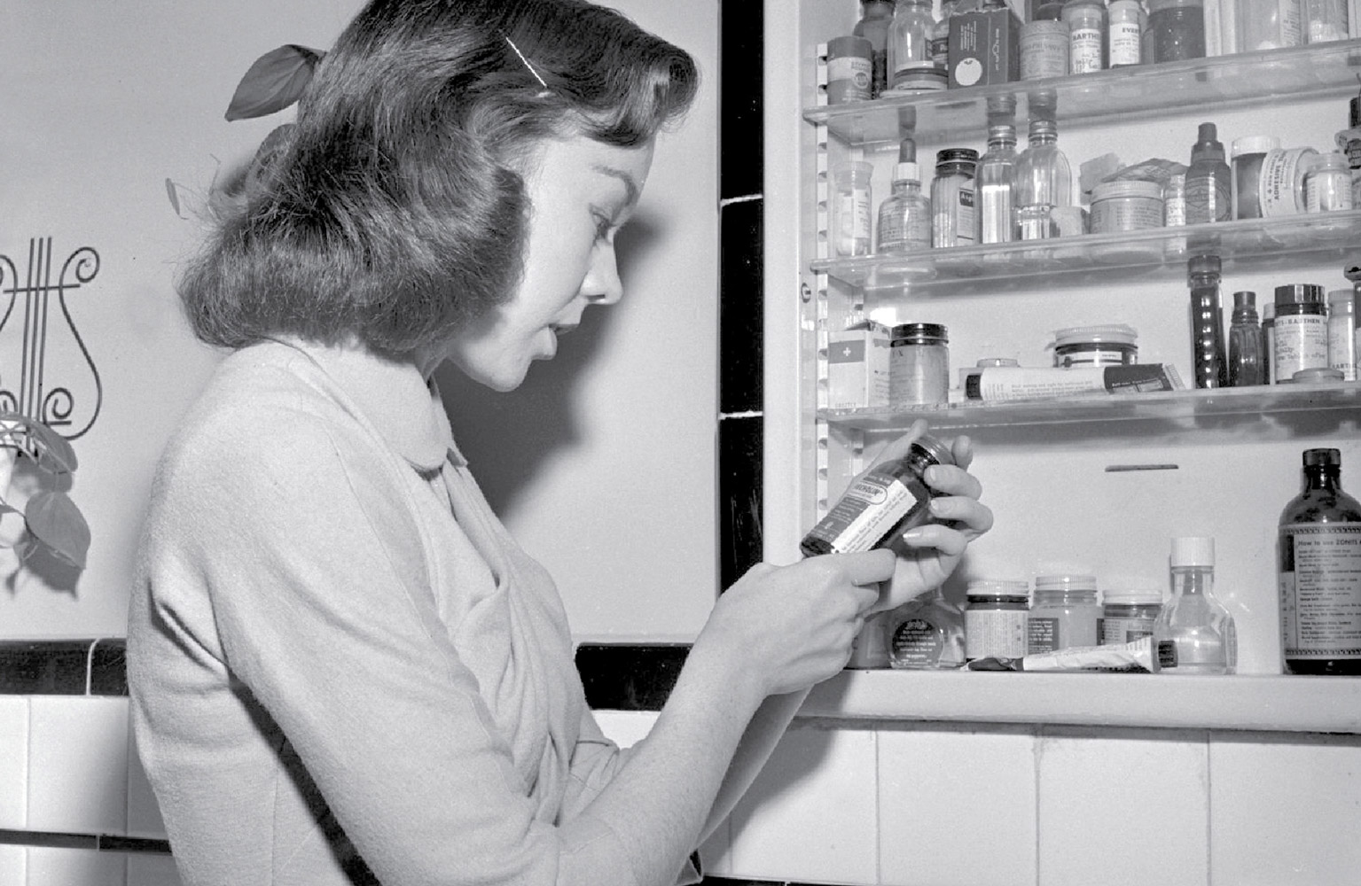 A photograph of a woman looking at a bottle from the medicine cabinet in a bathroom.