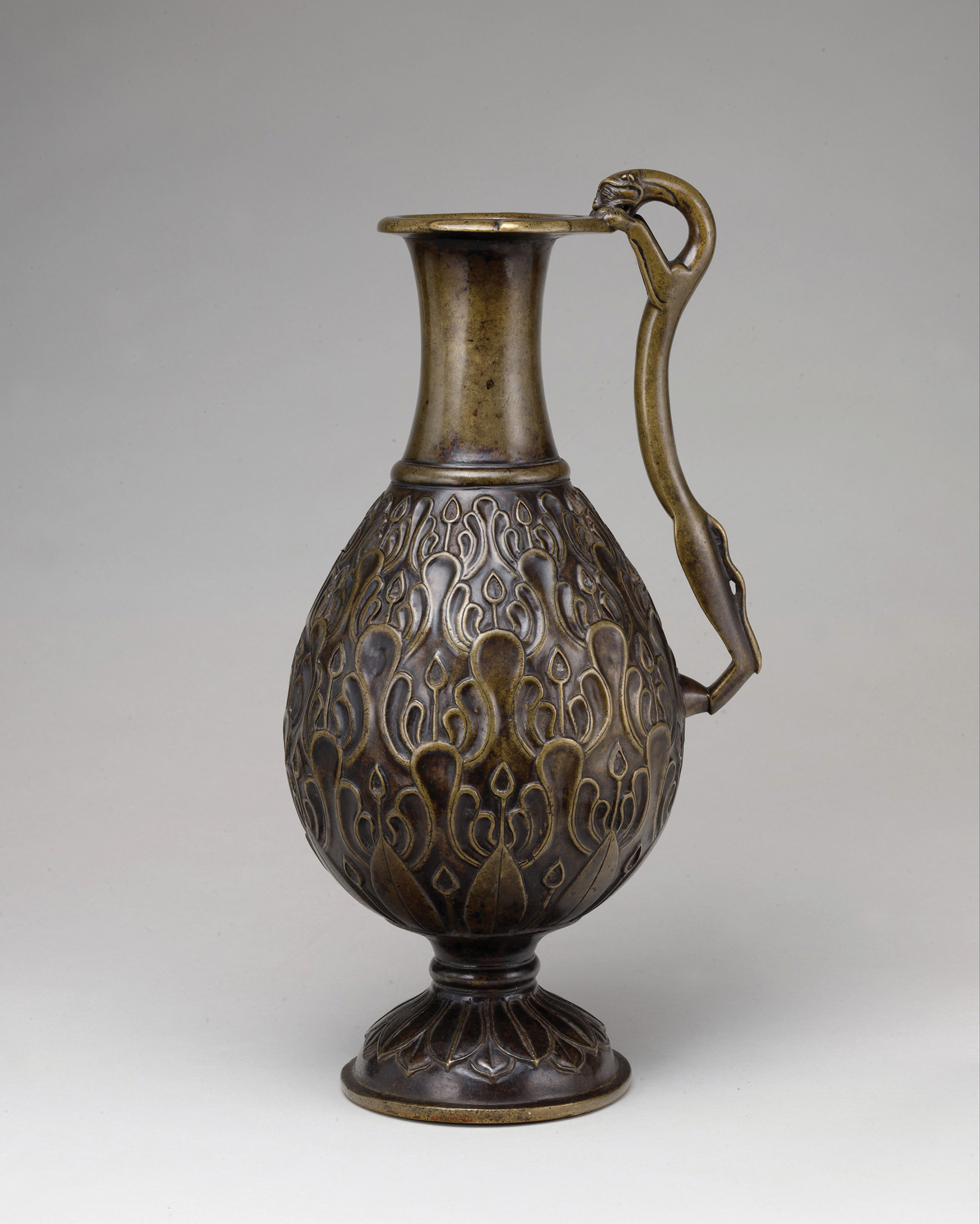A photograph of an Iranian ewer from the seventh century. The elongated cat that forms the handle is peering at the heads of two birds depicted on the rim of the vessel. 