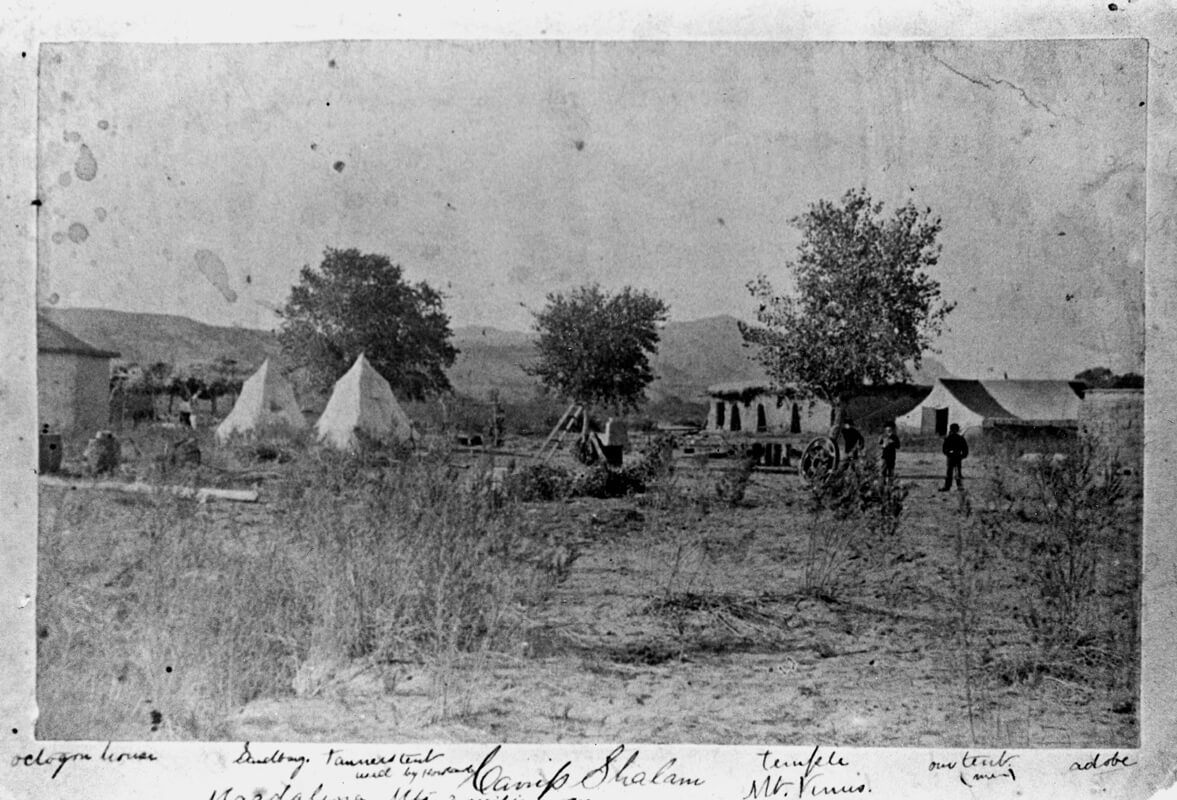 The earliest known photo of Shalam Colony, circa eighteen eighty five, depicting a collection of structures among a mountainous backdrop. The handwritten captions along the bottom margin identify both structures in the camp—such as “octogon house” and the “temple,” which was likely the first building erected at the site—as well as natural landmarks including Robledo Peak, known at the time as Mount Venus.
