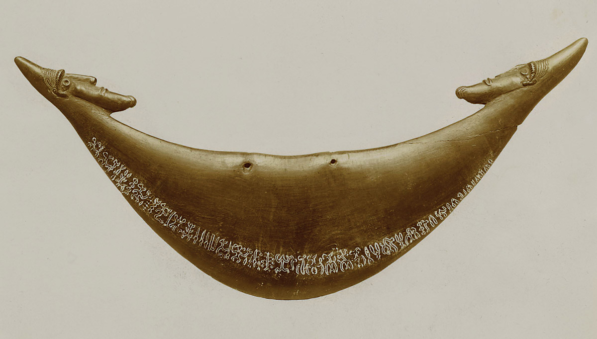 A photograph of a crescent-shaped, wooden neck ornament from Easter Island made some time in the first half of the nineteenth century. The artifact, decorated with two bearded male heads on either end, contains a line of rongorongo glyphs along its bottom edge. 