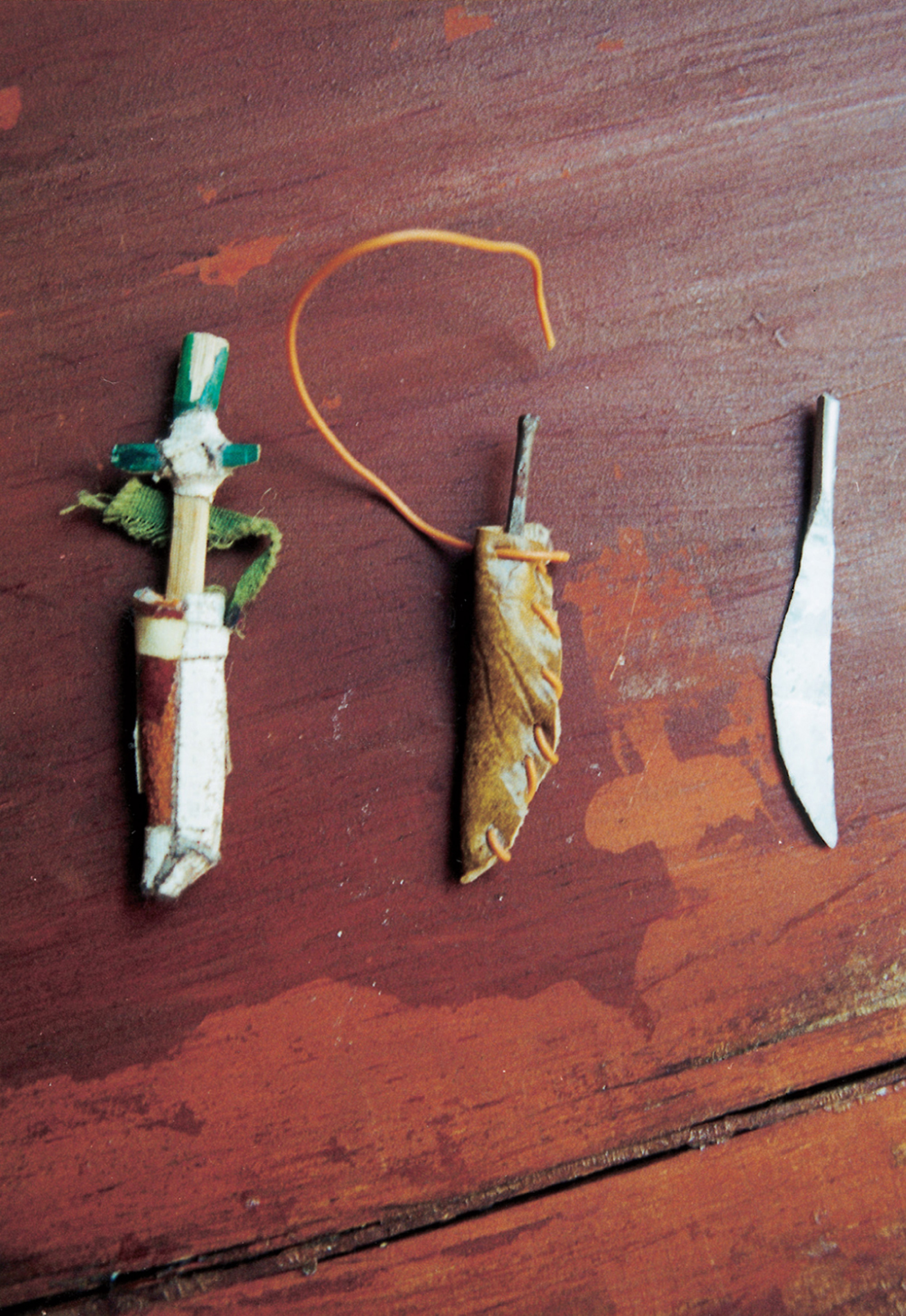 Lot of three daggers.  
1. Steel dagger made of a round-headed nail, half of which has been hammered flat to make a straight, narrow blade; the sheath is faux leather, folded and stitched up one side with telephone wire, the end of which is left loose to serve as a strap. 2. Aluminum dagger made of a length of wire hammered flat. 3. Bamboo dagger, whittled flat and tapered toward the tip. The grip is colored green on one side; a short hand-guard is affixed to the blade with white medical cloth tape. The blade fits snugly inside its sheath, which is real leather folded and joined with more cloth tape. A thin strip of fraying green cloth is taped to the top of the sheath; this once formed a loop for hanging, but is torn close to the sheath on one end. Daggers, not all of them as finely crafted as these, were de rigueur for the heroes and heroines (above all the spunky Aina) of the Doll Games’ “Pirate” and “Outlaw” scenarios.