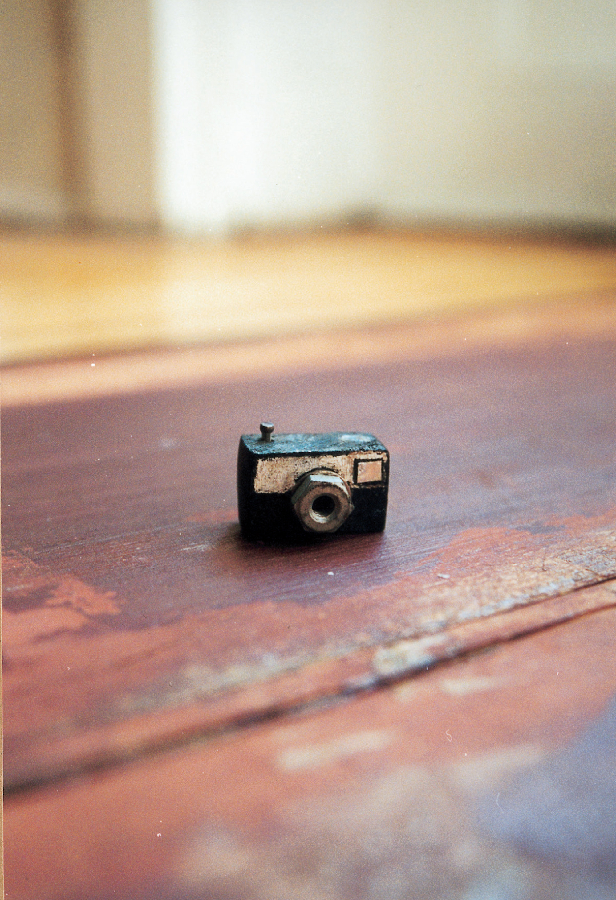 A photograph depicting a small crafted camera. The caption reads: Camera. Rectangular block of wood painted black with a white strip accenting the front, where a nut and washer together form the lens. The viewfinder window is indicated on this side by a small rectangle of silver metallic paper or tape inset in the white strip and outlined in black. On the top of the camera the head of a small nail driven most of the way into the body of the camera represents the shutter release. On the back of the camera a small strip of yellow paper is glued to the center. On it is printed “1—1” to represent the exposure number. This camera, loaded with self-reflexive implications (and a full roll of film), also points to the voyeurism/exhibitionism so characteristic of Doll Games plots.
