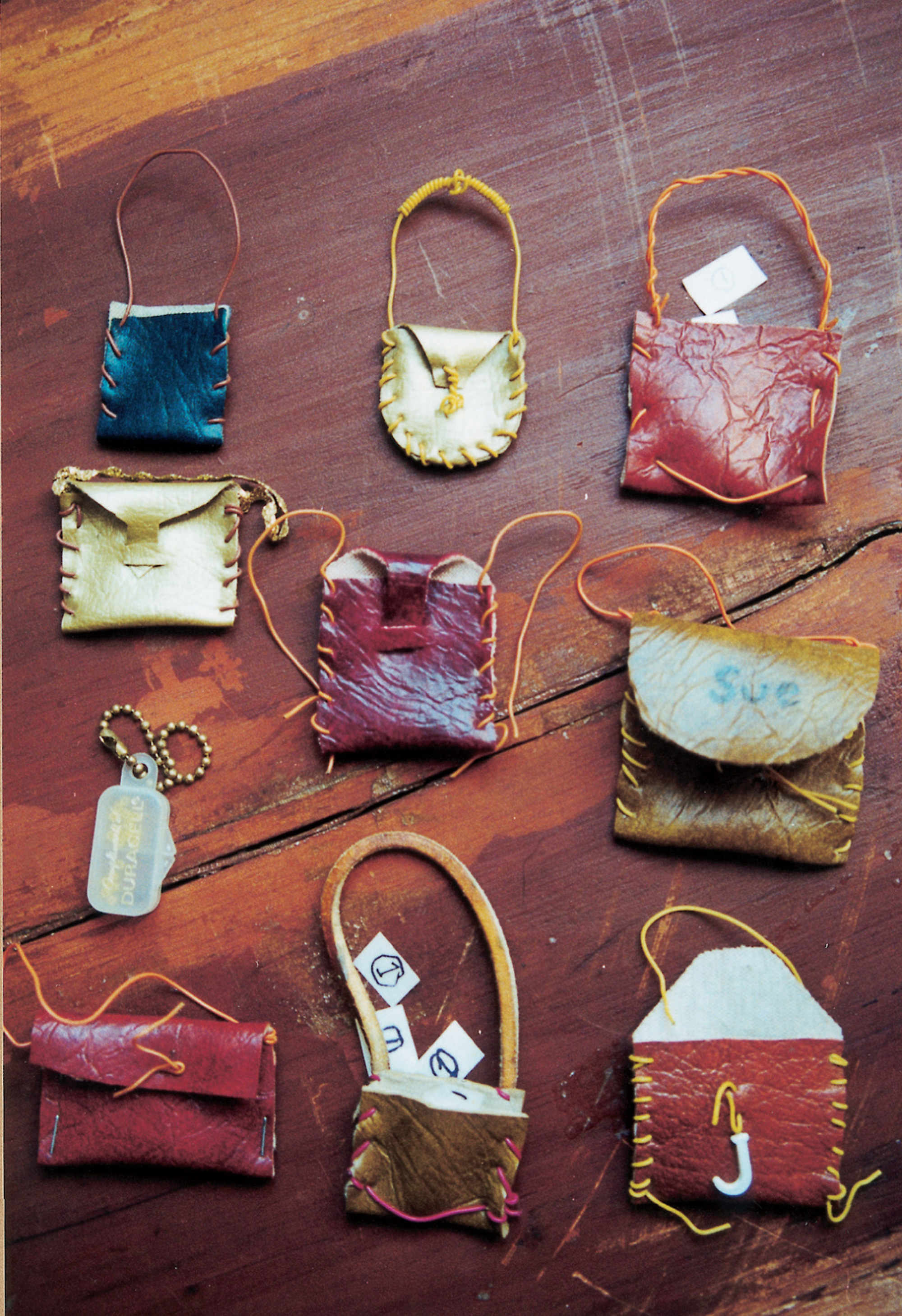 Eight assorted handbags, one purse and one backpack. The Duracell purse, semi-transparent purse on small brass bead-chain, was made from a container for hearing aid batteries. All others are of faux leather, most stitched with telephone wire. Two contain paper “money,” white rectangles each bearing a circled numeral 1 in pencil/black marker. Paradoxically, although the Doll Games showed a utopian disregard for money and a high scorn for the conventional appurtenances of femininity, purses were manufactured in quantities rivaled only by daggers. Was the vaginal purse—pictured here in an almost military array—waging a war with the phallic dagger over the contested territory of the Doll Games?